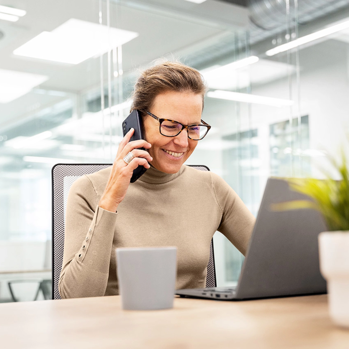 Eine Frau sitzt lächelnd in einem Büro vorm Laptop und telefoniert mit dem Handy