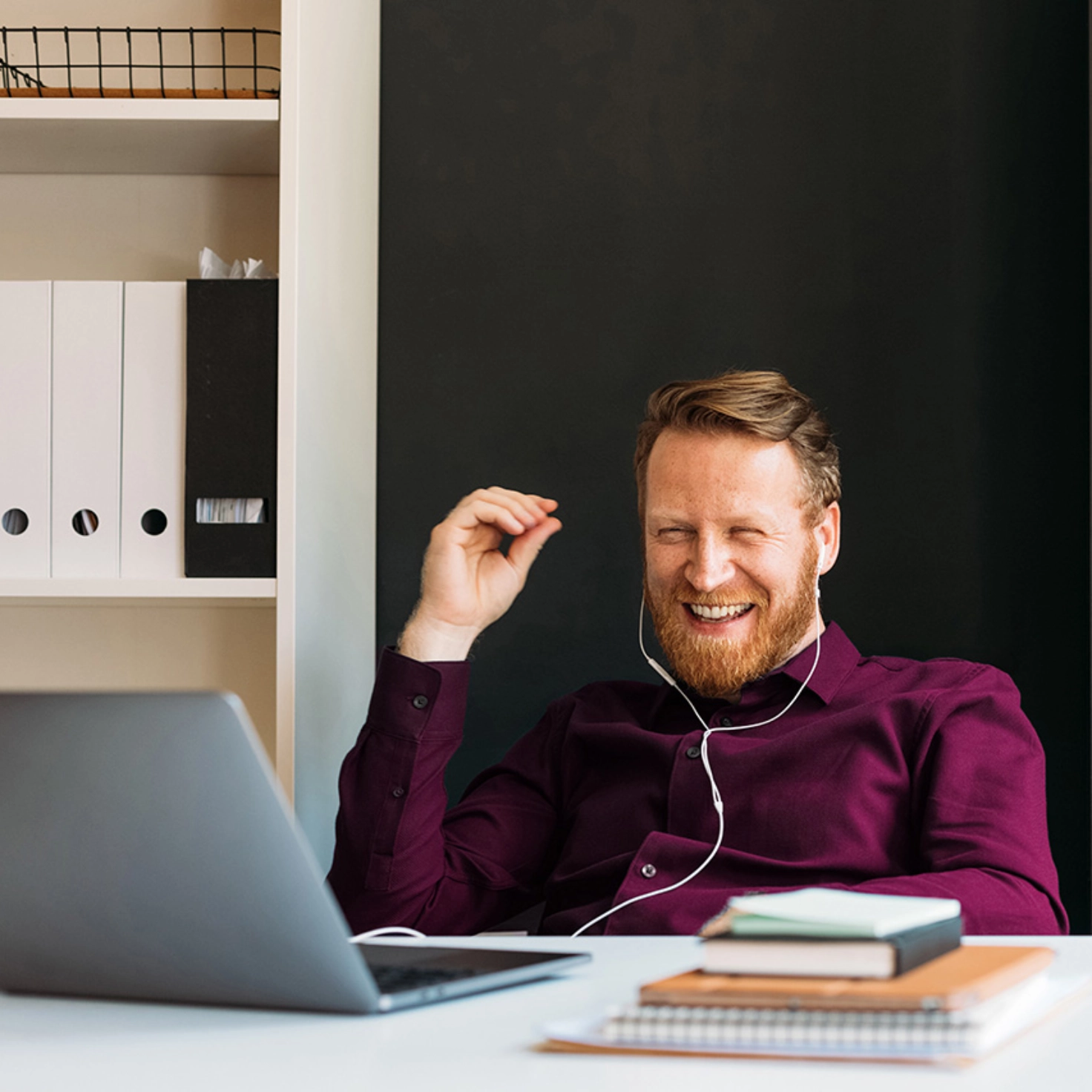 Ein Mann mit roten Haaren, Bart und weinrotem Hemd sitzt mit weissen In-ear-Kopfhörern im Homeoffice und lacht während eines Videocalls in die Laptop-Kamera.