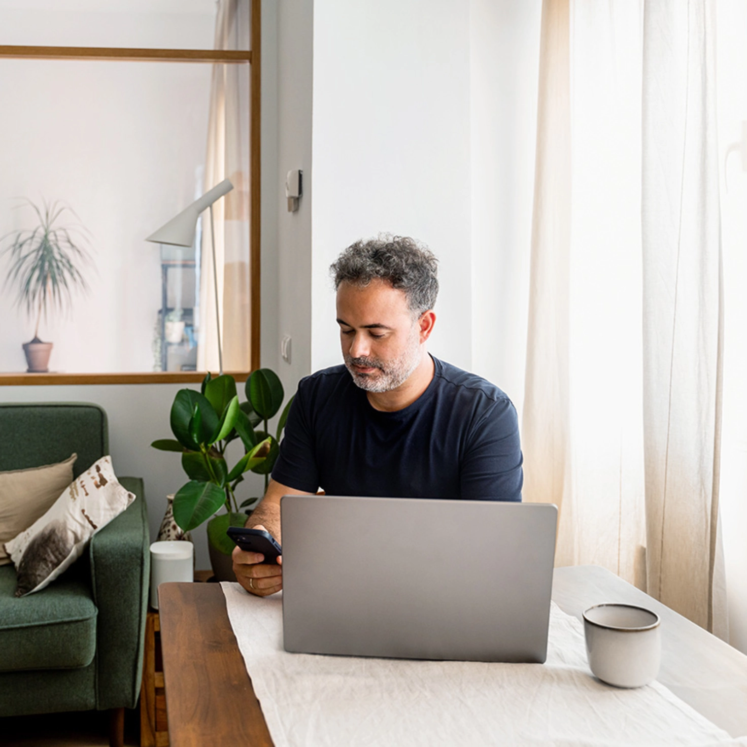 Ein Mann sitzt mit dem Smartphone in der Hand vor seinem aufgeklappten Laptop