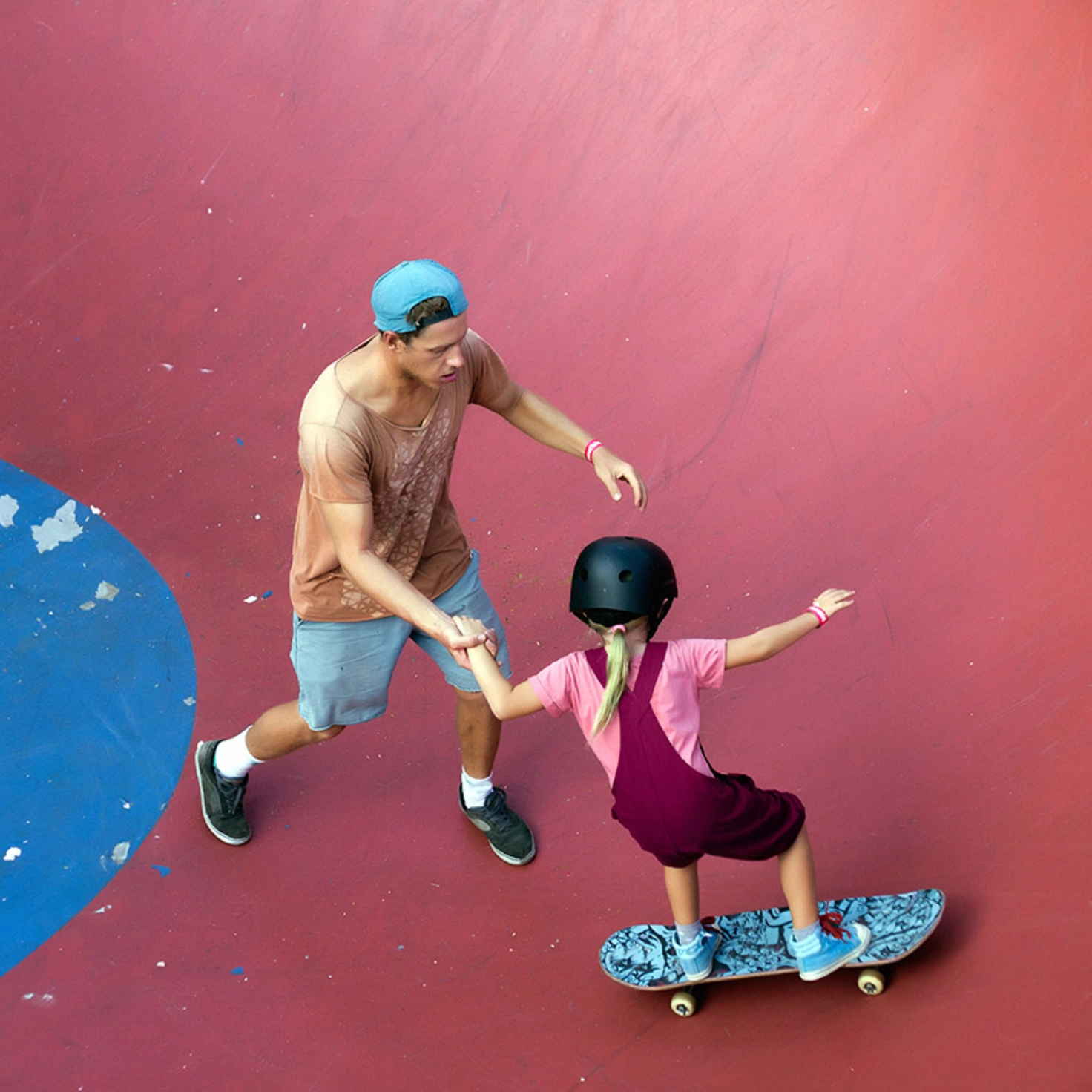 Ein Mädchen fährt auf dem Skateboard in einem Skatepark. Ihr Vater hält sie an der Hand.