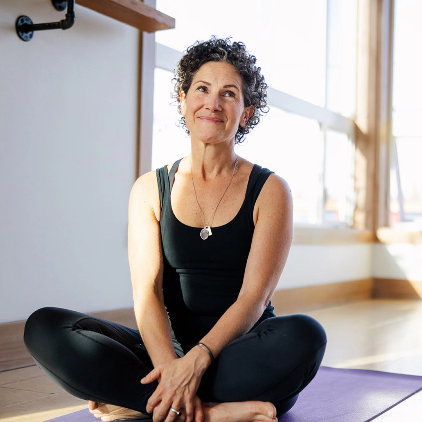 Eine Frau mit kurzen, lockigen Haaren, schwarzem Shirt und schwarzer Hose sitzt im Schneidersitz auf einer Yogamatte