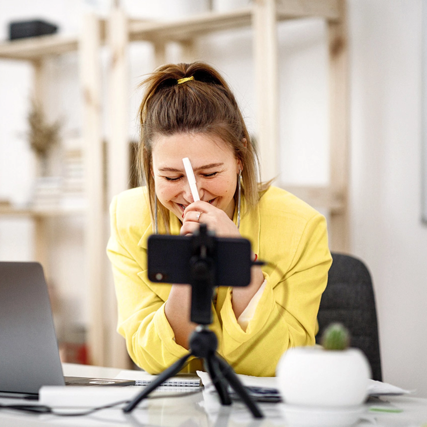 Eine junge Frau sitzt im Homeoffice am Tisch, den aufgeklappten Laptop vor sich und das Handy auf dem Stativ. Sie führt gerade einen Videocall am Handy und lacht dabei.