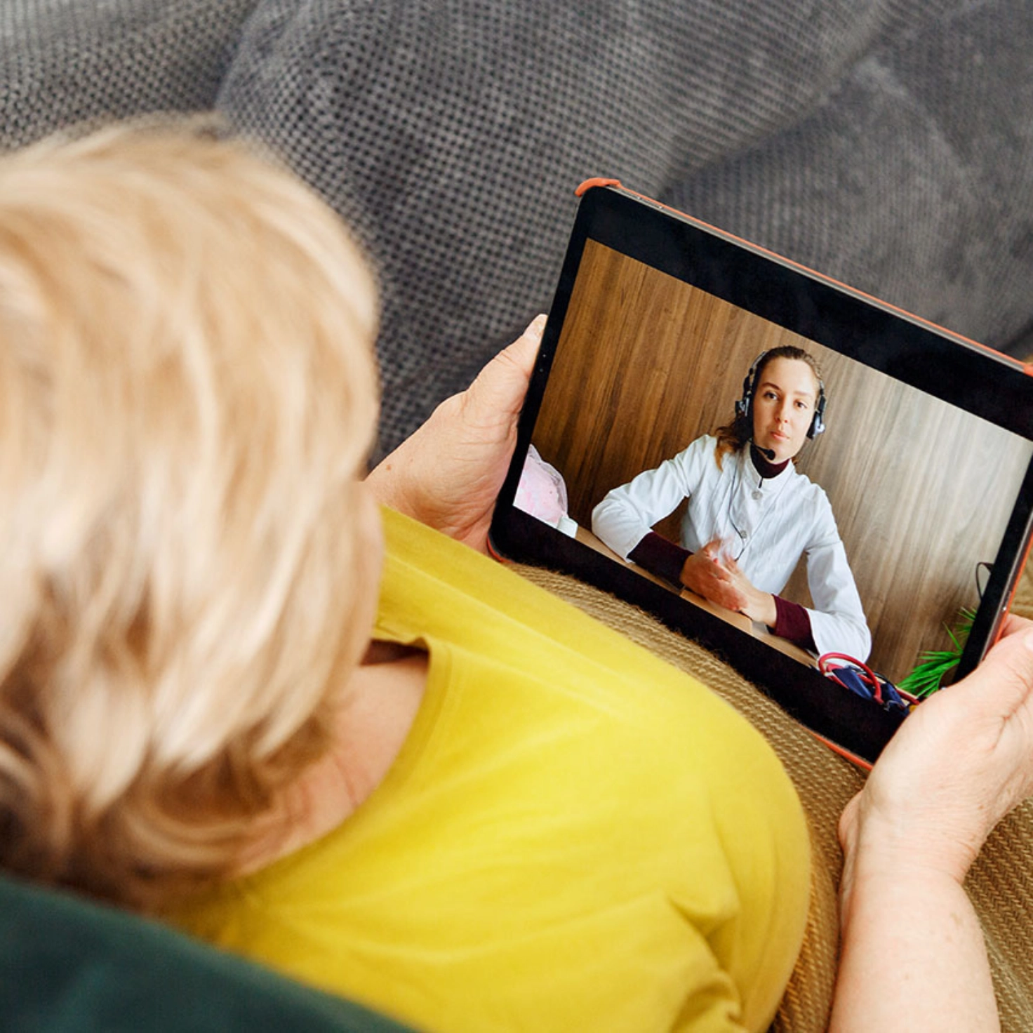 Eine Frau sitzt zugedeckt auf dem Sofa und führt einen Videoanruf mit einer Ärztin am Tablet