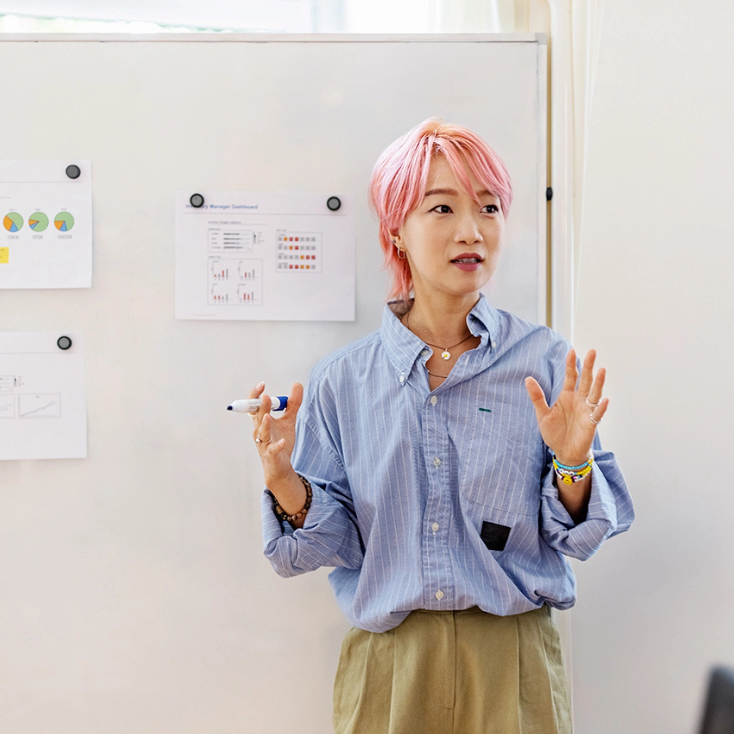 Eine junge Frau mit rosa Haaren steht vor einem Flipchart, an dem mit Magneten einige Dokumente hängen, und hält eine Präsentation.