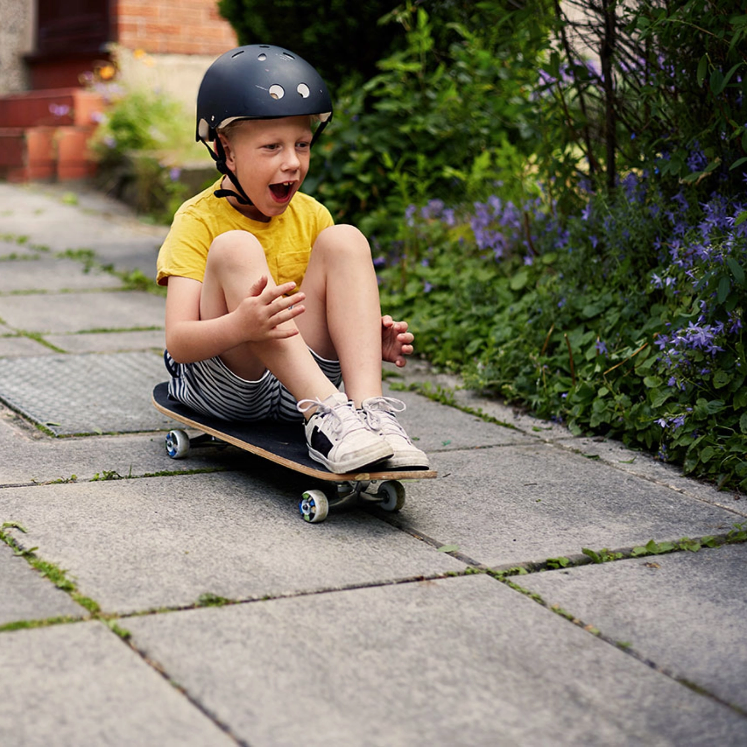 Ein Kleinkind mit Helm sitzt mit angewinkelten Beinen auf einem Skateboard und fährt lachend den Weg lang