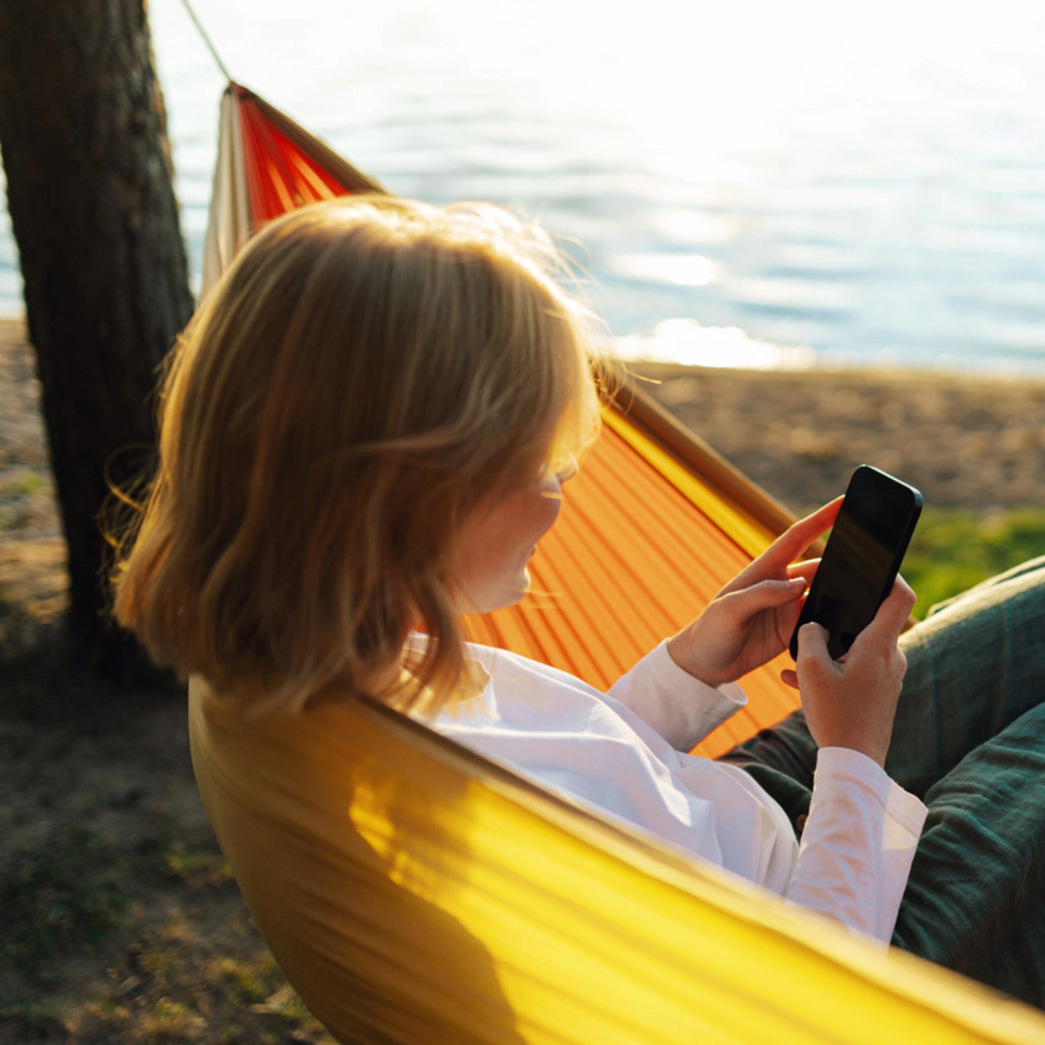 Eine Frau sitzt in einer gelben Hängematte an einem See und schaut auf ihr Handy