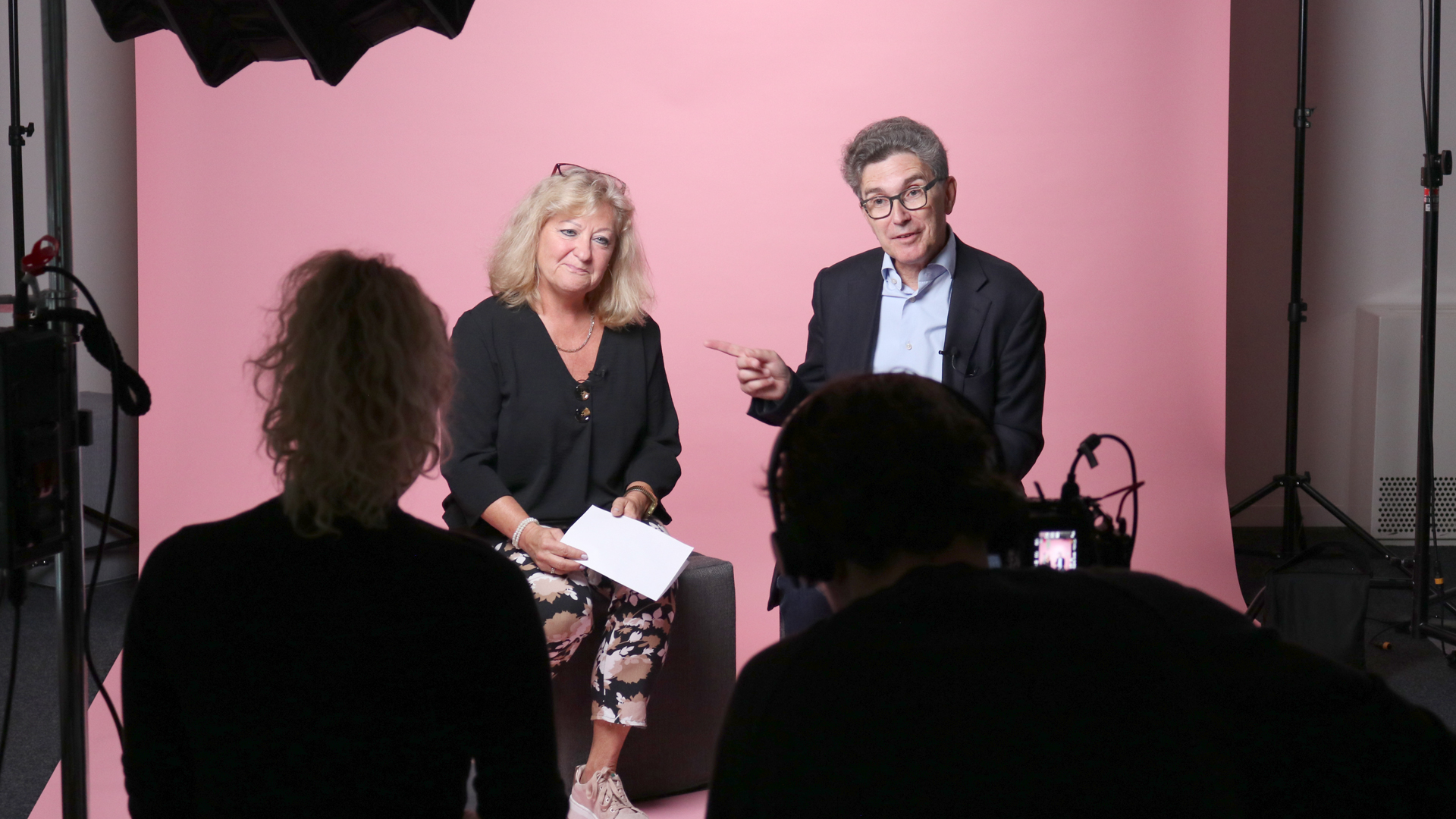 Snapshot from the video shoot with the Sympany Customer Council: The focus is on a man and a woman sitting in front of a pink screen. The man speaks and gesticulates. The camera team can be seen from behind in the foreground.