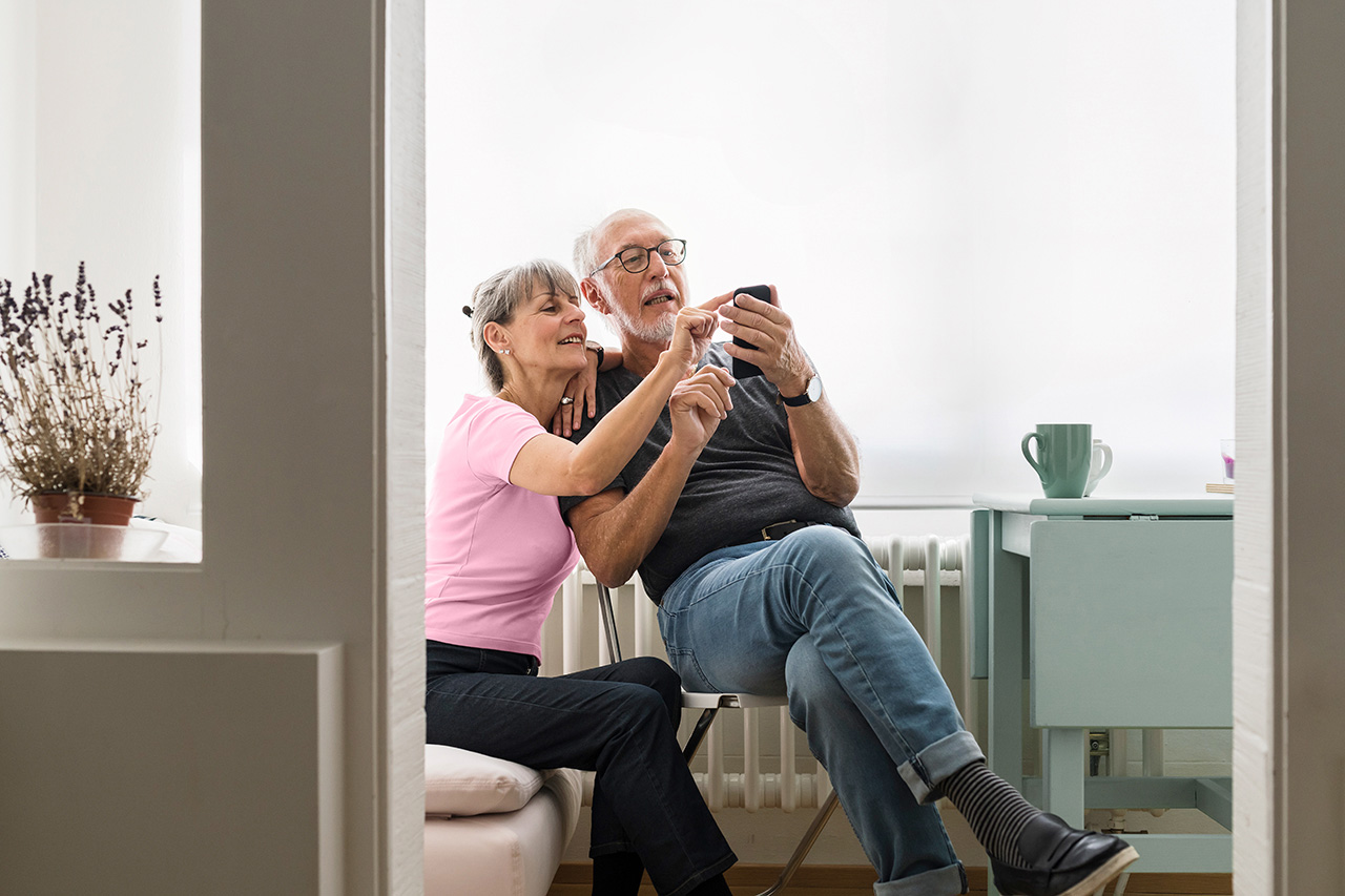 An elderly couple are sitting in the living room and typing on a mobile phone together.