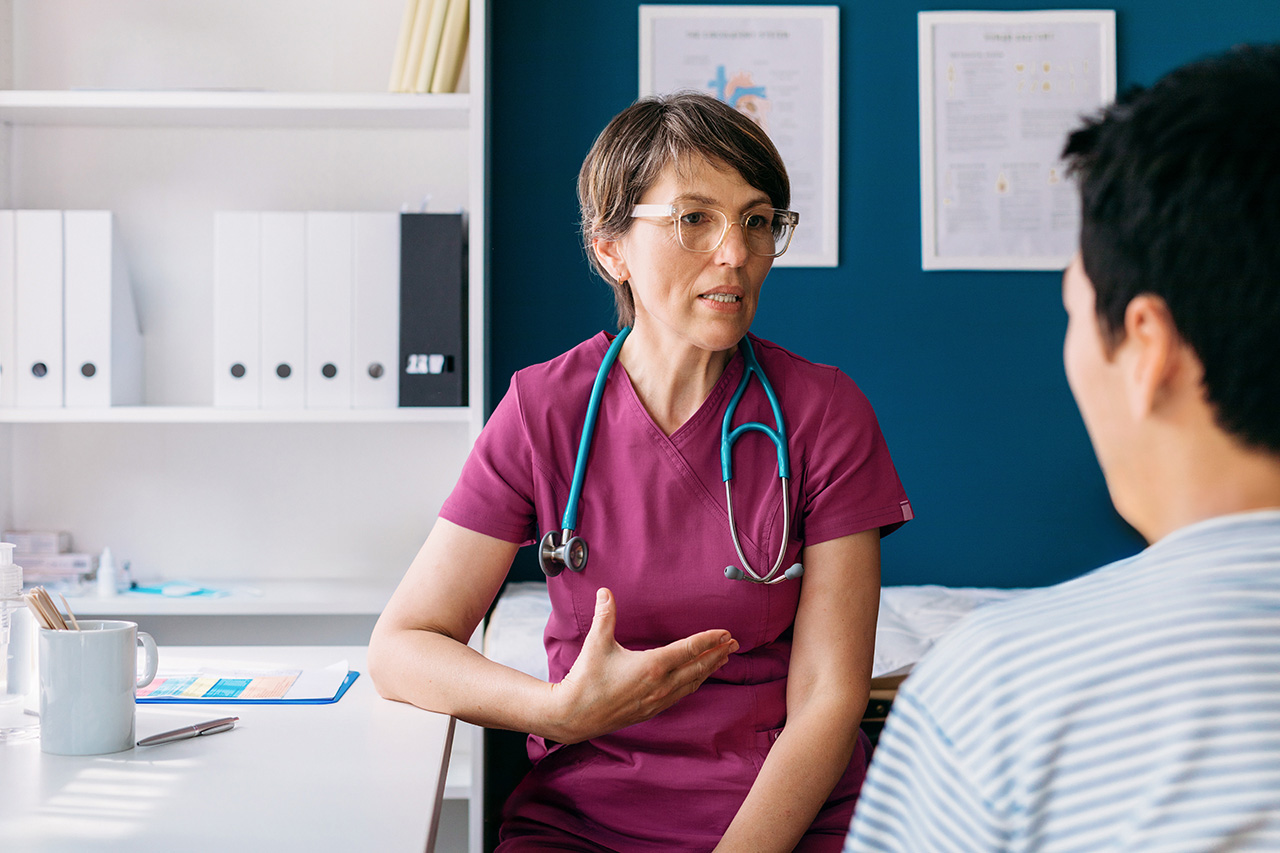 Eine Ärztin mit weinrotem Kittel und Stethoskop um den Hals führt ein Gespräch mit einem Patienten, der von hinten gezeigt wird