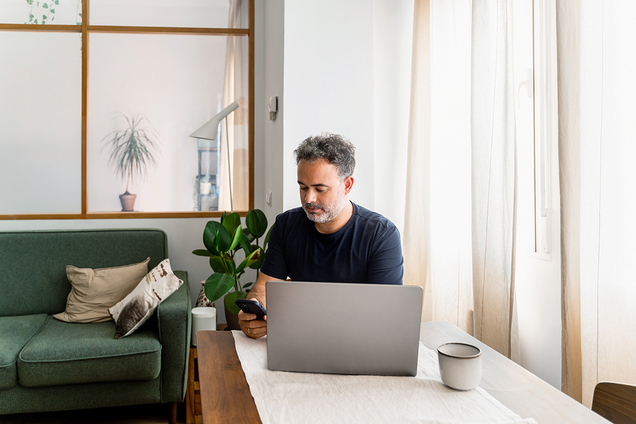 Ein Mann sitzt mit dem Smartphone in der Hand vor seinem aufgeklappten Laptop