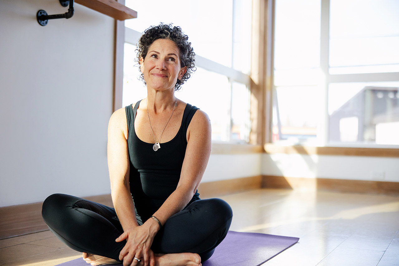 Eine Frau mit kurzen, lockigen Haaren, schwarzem Shirt und schwarzer Hose sitzt im Schneidersitz auf einer Yogamatte