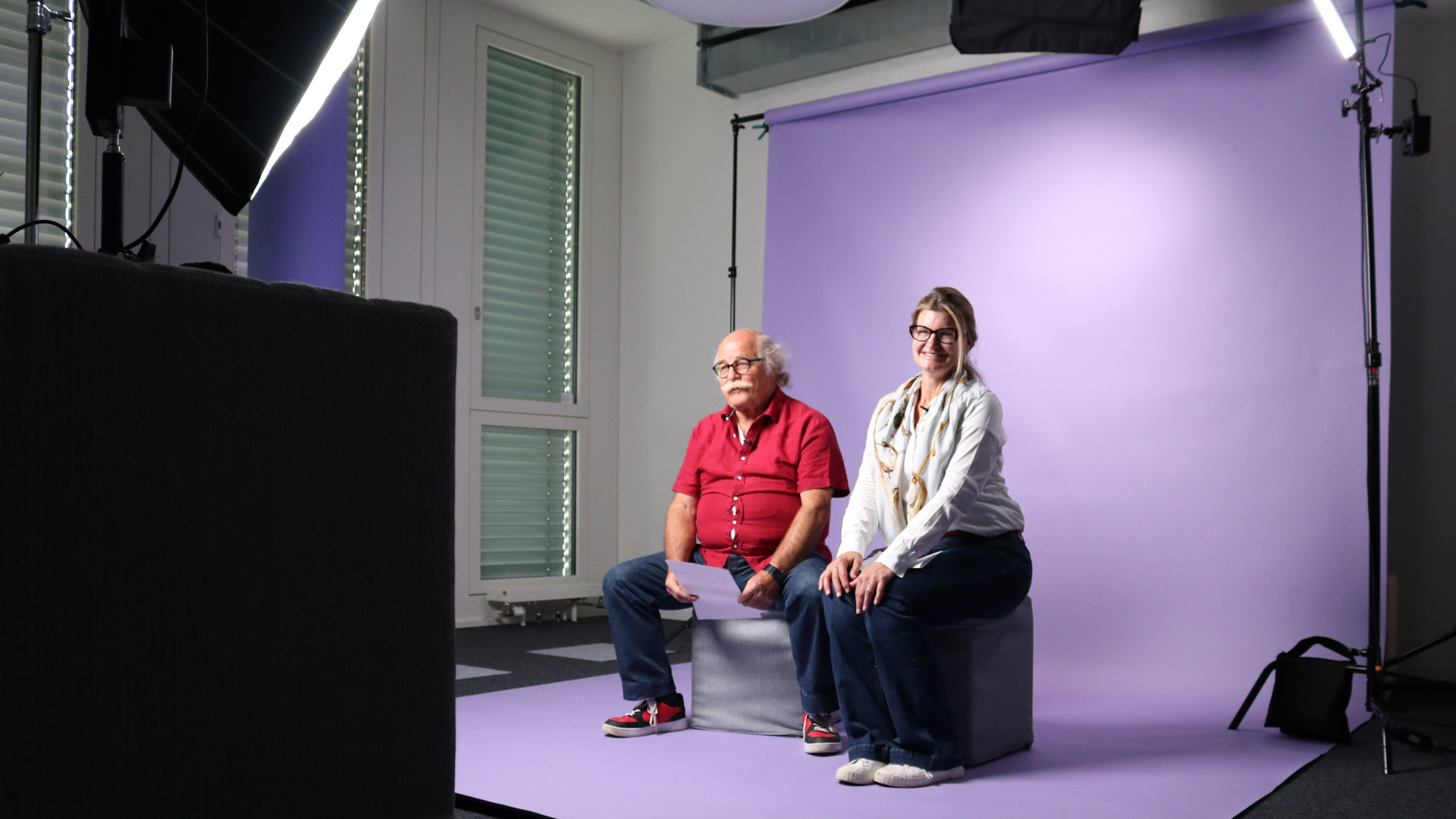 Snapshot from the video shoot with the Sympany Customer Council: A man and a woman sit on cubes in front of a purple canvas