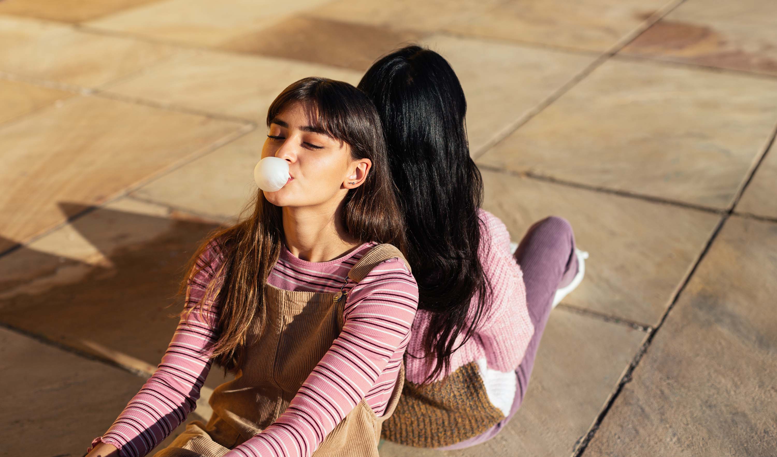 Two women are sitting back to back on the floor with their legs drawn up. The woman looking at the camera makes a big bubble gum bubble.